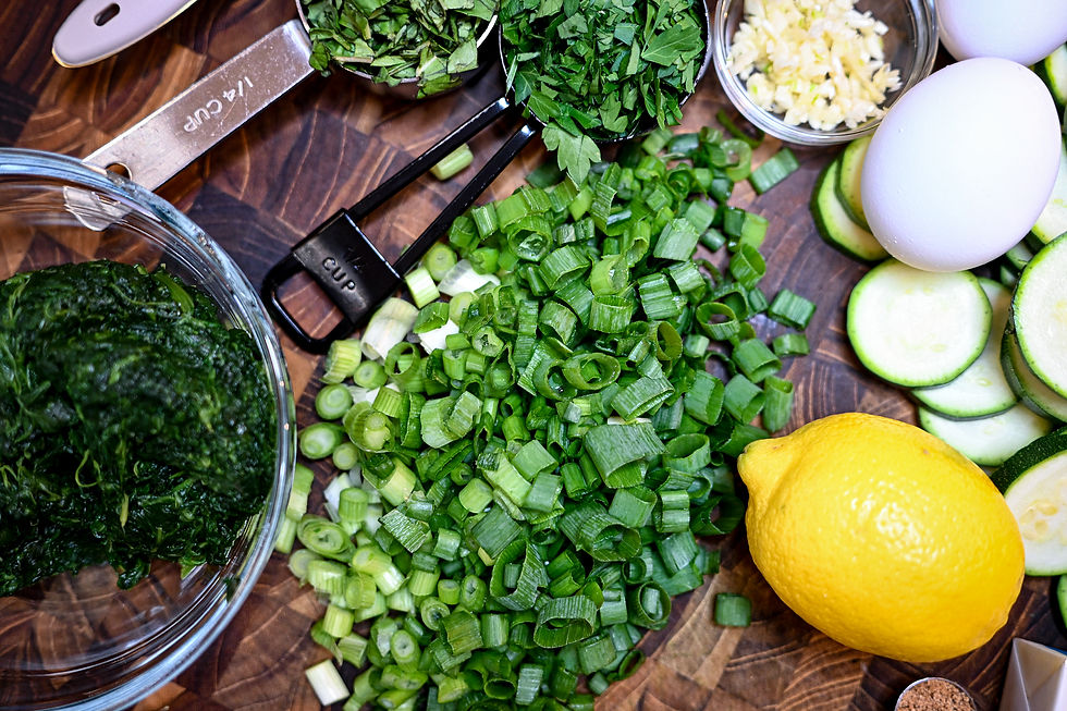 Chopped green onions, spinach, herbs, sliced zucchini, lemon, eggs, and garlic on a wooden board with measuring cups. Bright and fresh.