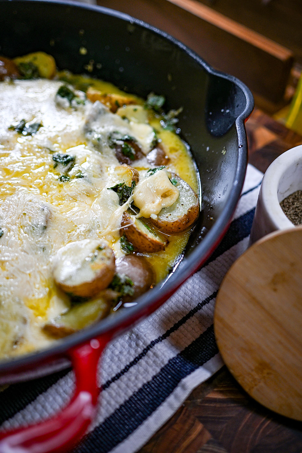 Cheesy skillet meal with sliced sausage, spinach, and melted cheese in a red pan on a striped cloth, next to a wooden board.