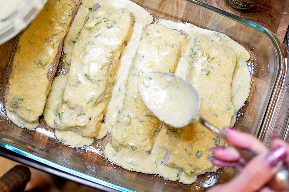Glass dish with fish fillets covered in mustard-green sauce. Close-up of hand using a spoon to spread sauce. Wooden table.