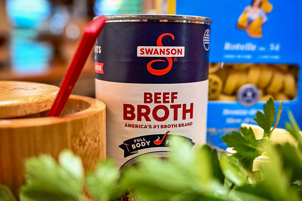 Swanson beef broth can beside a red spoon and herbs in a kitchen, with a blue pasta box blurred behind.
