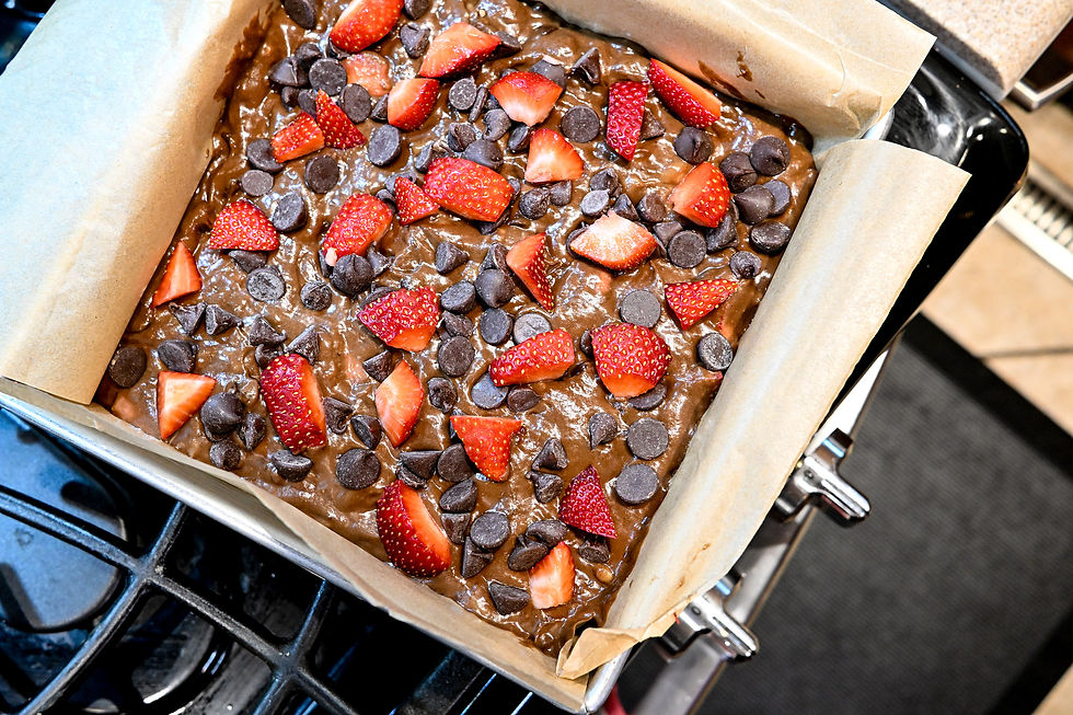 Brownie batter in a parchment-lined pan topped with fresh strawberry slices and chocolate chips, placed on a stove, ready to bake.
