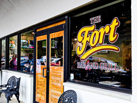 The image shows The Fort Bar and Grill's entrance with a large sign on the window. Outdoor seating includes decorative metal chairs.