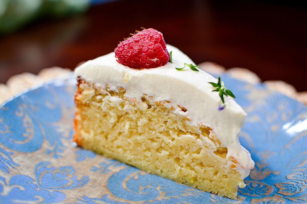 Slice of lemon cake with white frosting and a raspberry on top, on a blue decorative plate. Background is softly blurred.