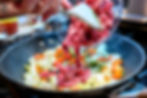 Raw beef being tipped into a wok with onions, carrots, and peppers on a stovetop, mid-cook in a busy kitchen