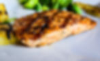 Grilled salmon garnished with herbs, golden brown and juicy, on a plate with grilled pineapple and broccoli in the background.