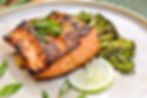 Grilled salmon garnished with green onions and sesame seeds, served on a plate with broccoli and a lime slice. Brown and green tones.