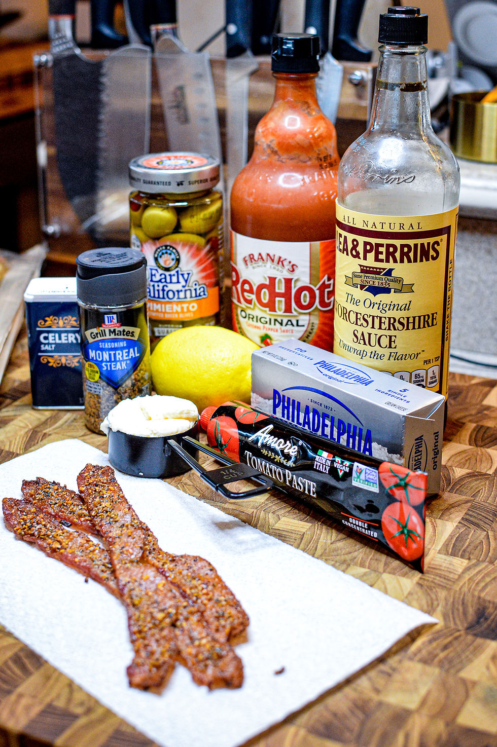 Ingredients on a wooden board: bacon, cheese, lemon, sauces, and spices, including Worcestershire, celery salt, and hot sauce.