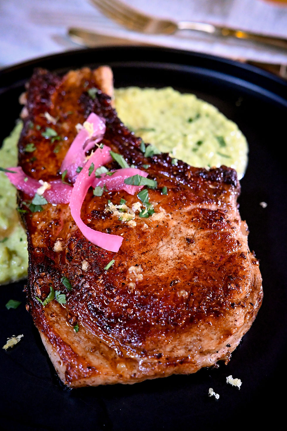 Grilled pork chop topped with pink pickled onions and herbs on a black plate, beside green sauce. Warm, appetizing setting.