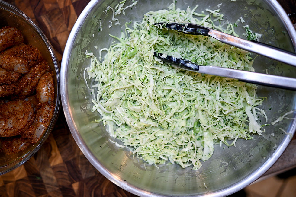 Shredded cabbage in a metal bowl with tongs, alongside seasoned shrimp on a wooden surface. Fresh, earthy tones dominate the scene.