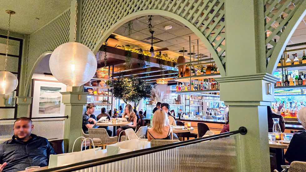 Restaurant scene with people dining under soft lighting. Green decor, bar in background, hanging plants, and spherical lamps create a cozy vibe.
