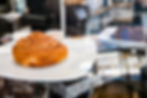 A cinnamon roll on a white plate with icing cups, next to a dark pastry in a cafe setting with a reflective surface.