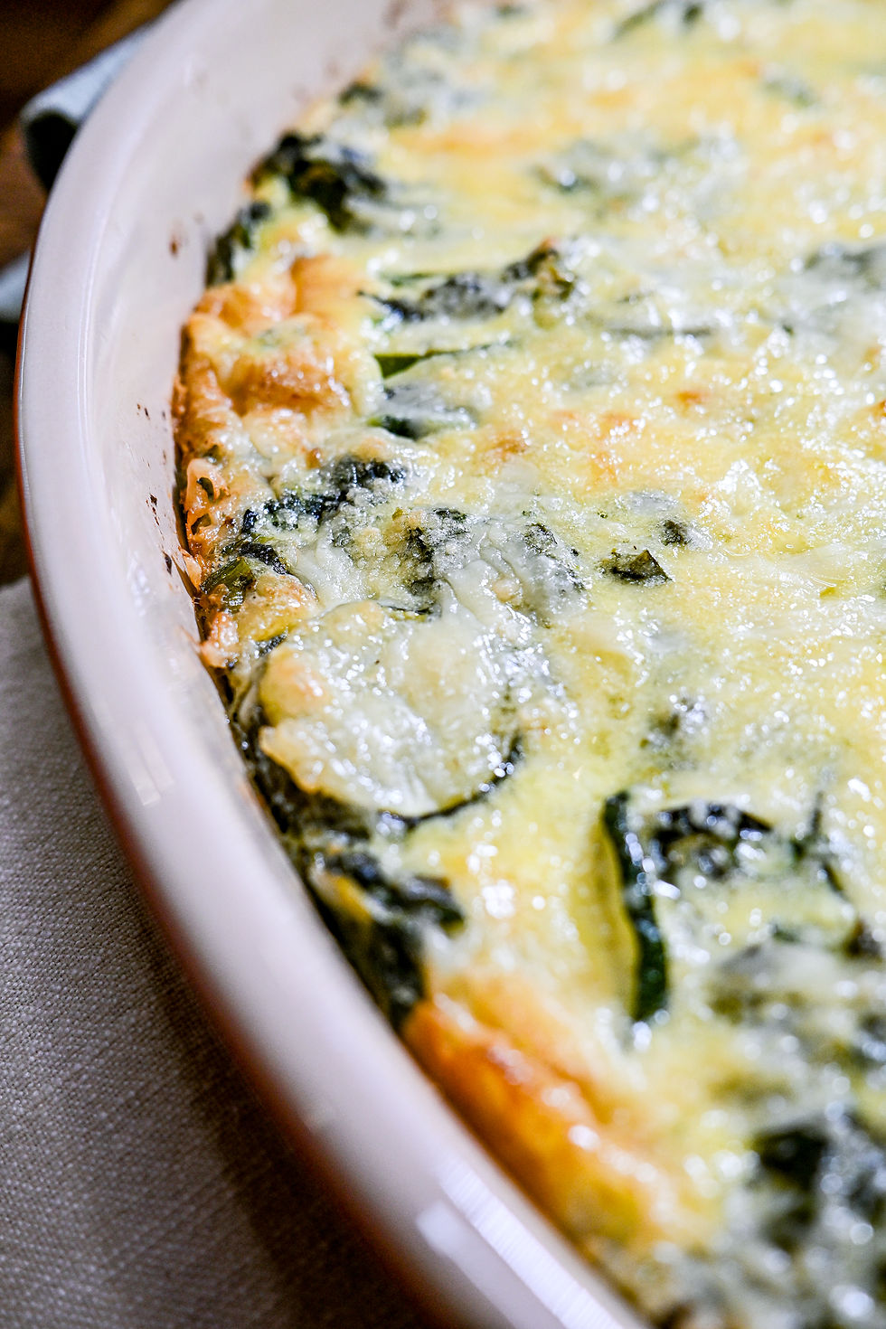 Close-up of a baked spinach casserole with golden cheese topping, in a white dish on a beige cloth. Mood: warm and appetizing.