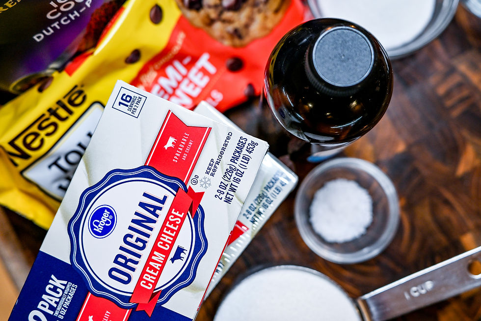 Cream cheese package, chocolate chips, and sugar on a wooden surface. Baking items include a vanilla bottle and measuring cup.