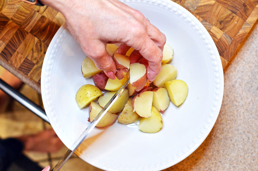 A hand cuts boiled potatoes in a white bowl on a wooden cutting board, with a knife. The setting is a kitchen countertop.