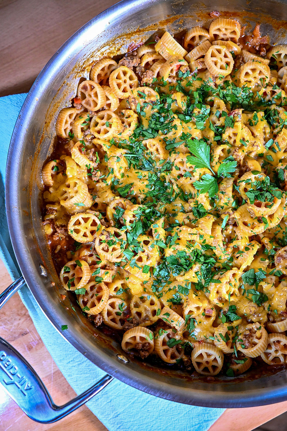 Skillet of baked wheel pasta with melted cheese, meat sauce, and chopped parsley on a wooden table.