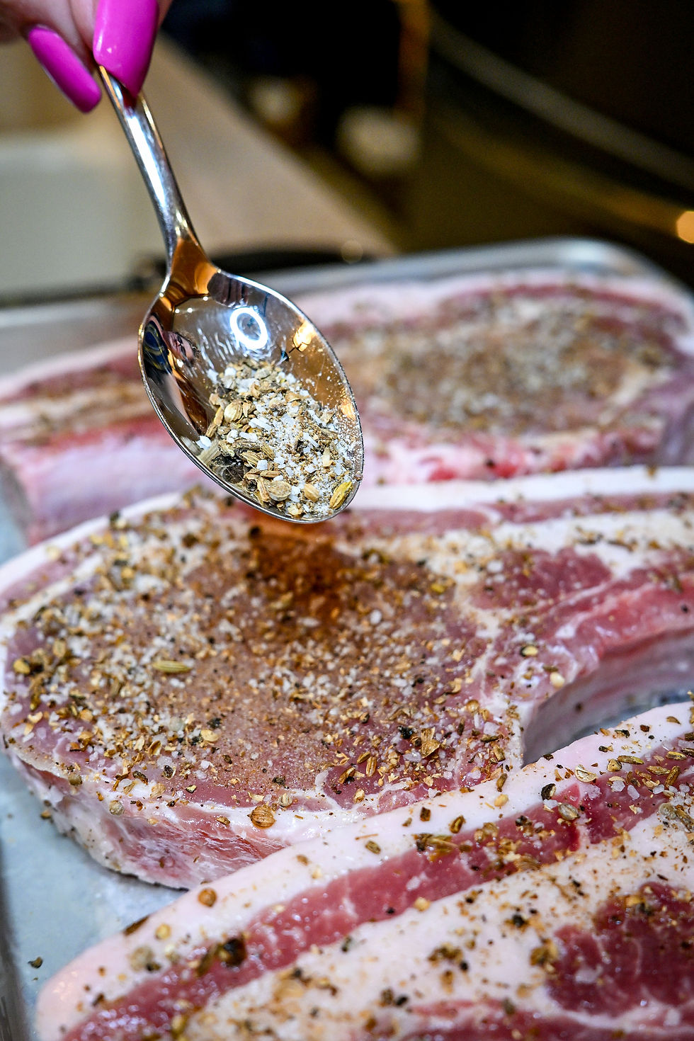 Hand with pink nails sprinkles spices from a spoon onto raw seasoned meat on a tray, in a kitchen setting.
