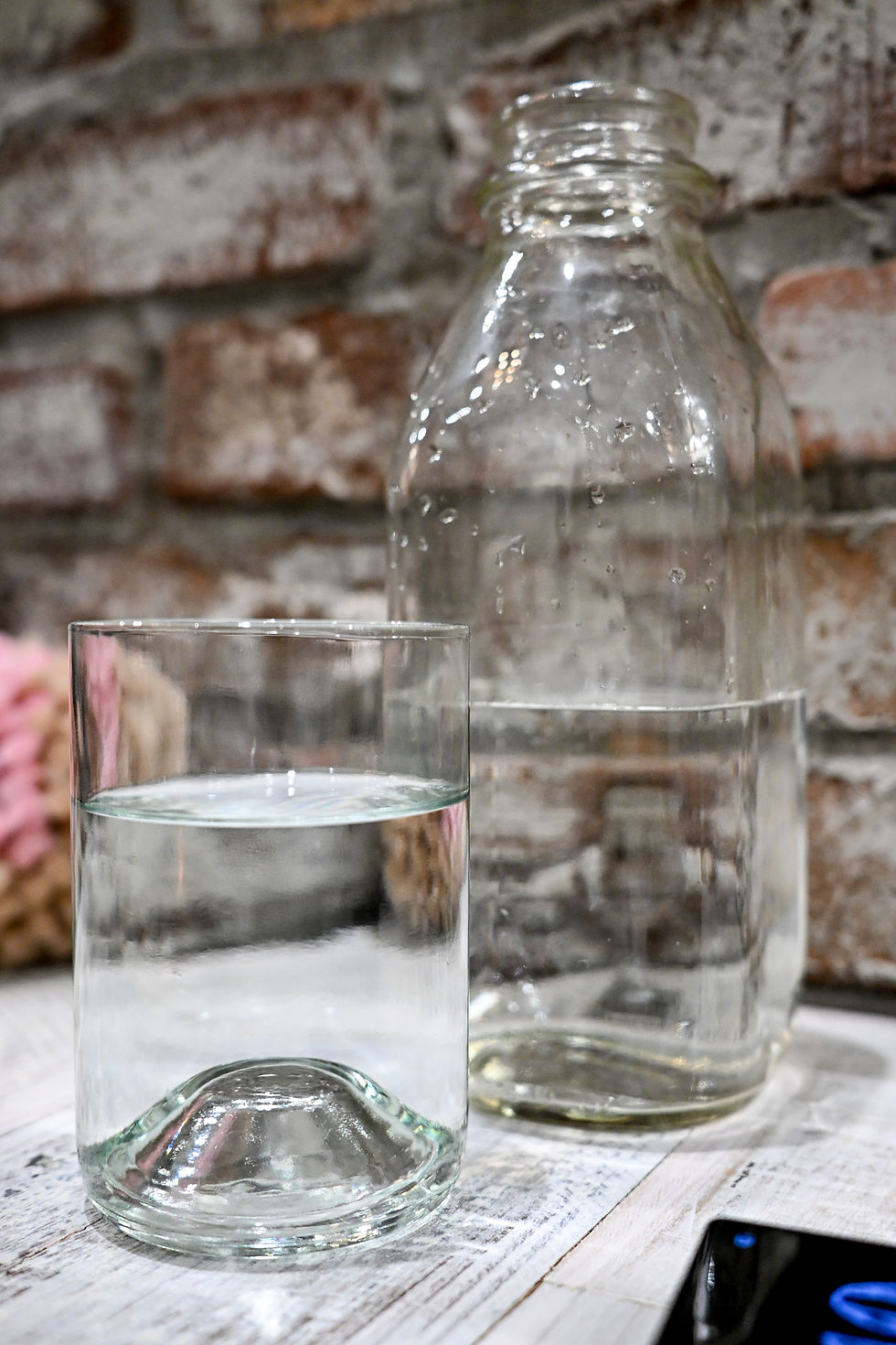 A glass and a bottle of water on a wooden table, with a rustic brick wall in the background. Soft, natural lighting.