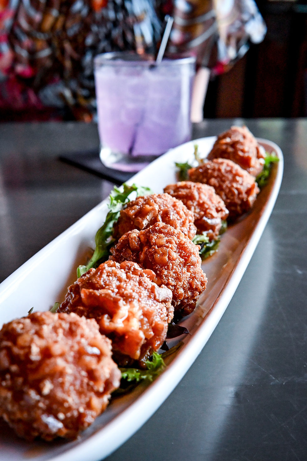 Plate of crispy fried chicken pieces on lettuce with a purple drink in a glass. Background has a colorful patterned fabric.