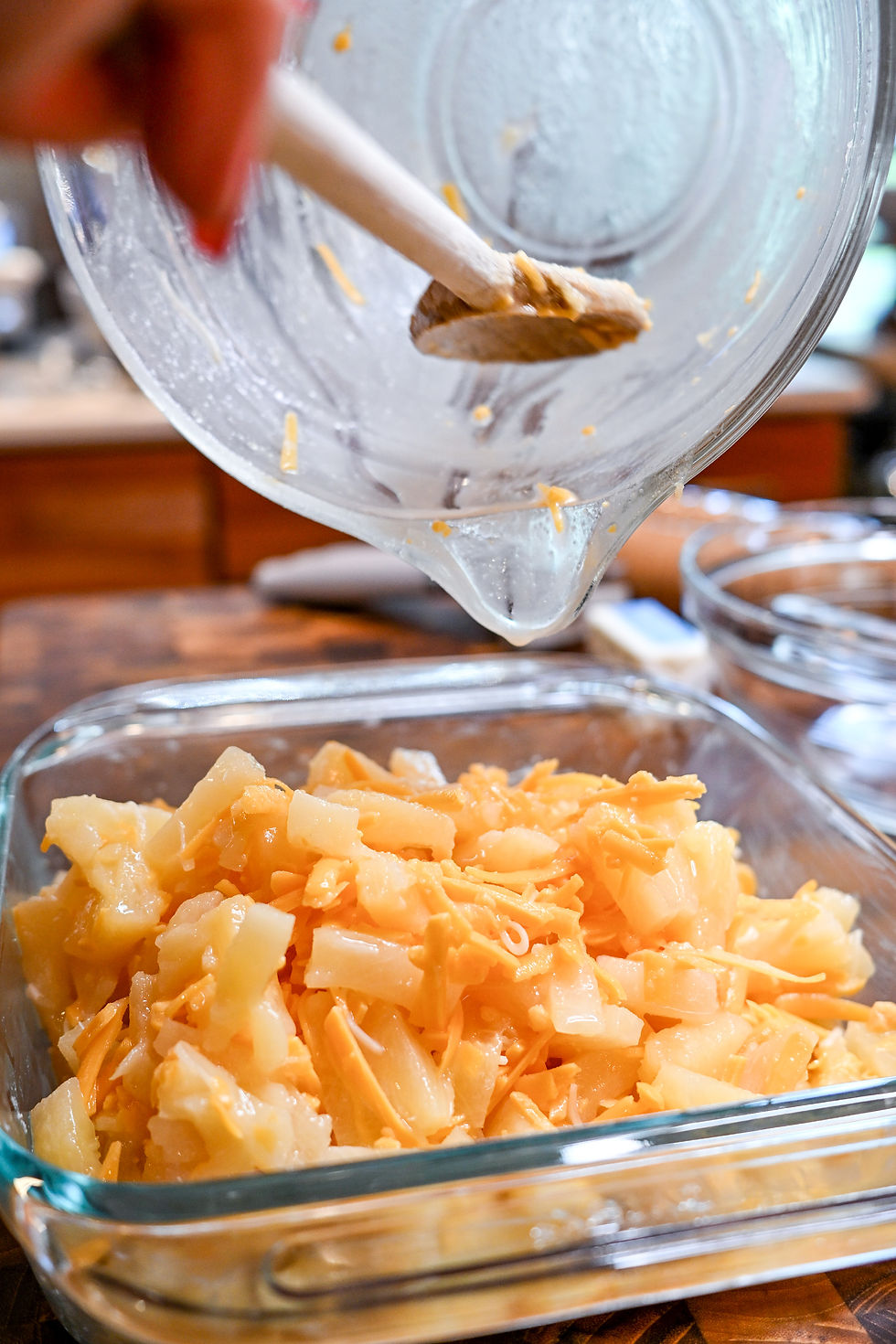 Hand pours shredded cheese and diced pineapple from a glass bowl into a baking dish in a kitchen setting. Warm, inviting mood.