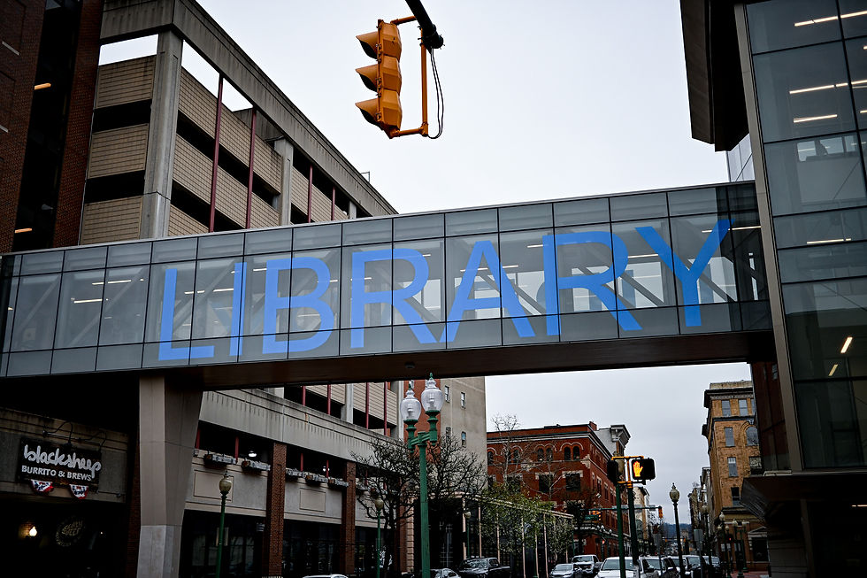 Glass walkway with large blue "LIBRARY" text connects buildings above a street. Visible traffic light and urban setting in the background.