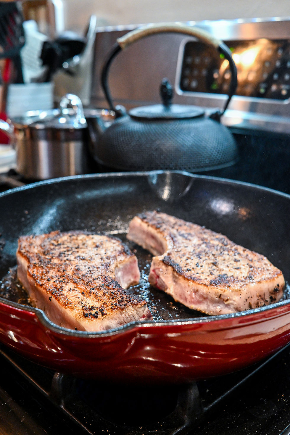 Two seasoned pork chops are sizzling in a red skillet on a stove. A black kettle and silver pot are in the blurred kitchen background.