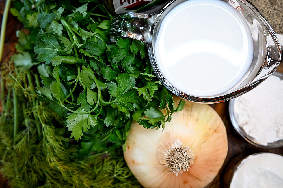 Fresh parsley, an onion, and dill surround a glass measuring cup filled with milk. Flour is visible near the edge. Bright, fresh scene.