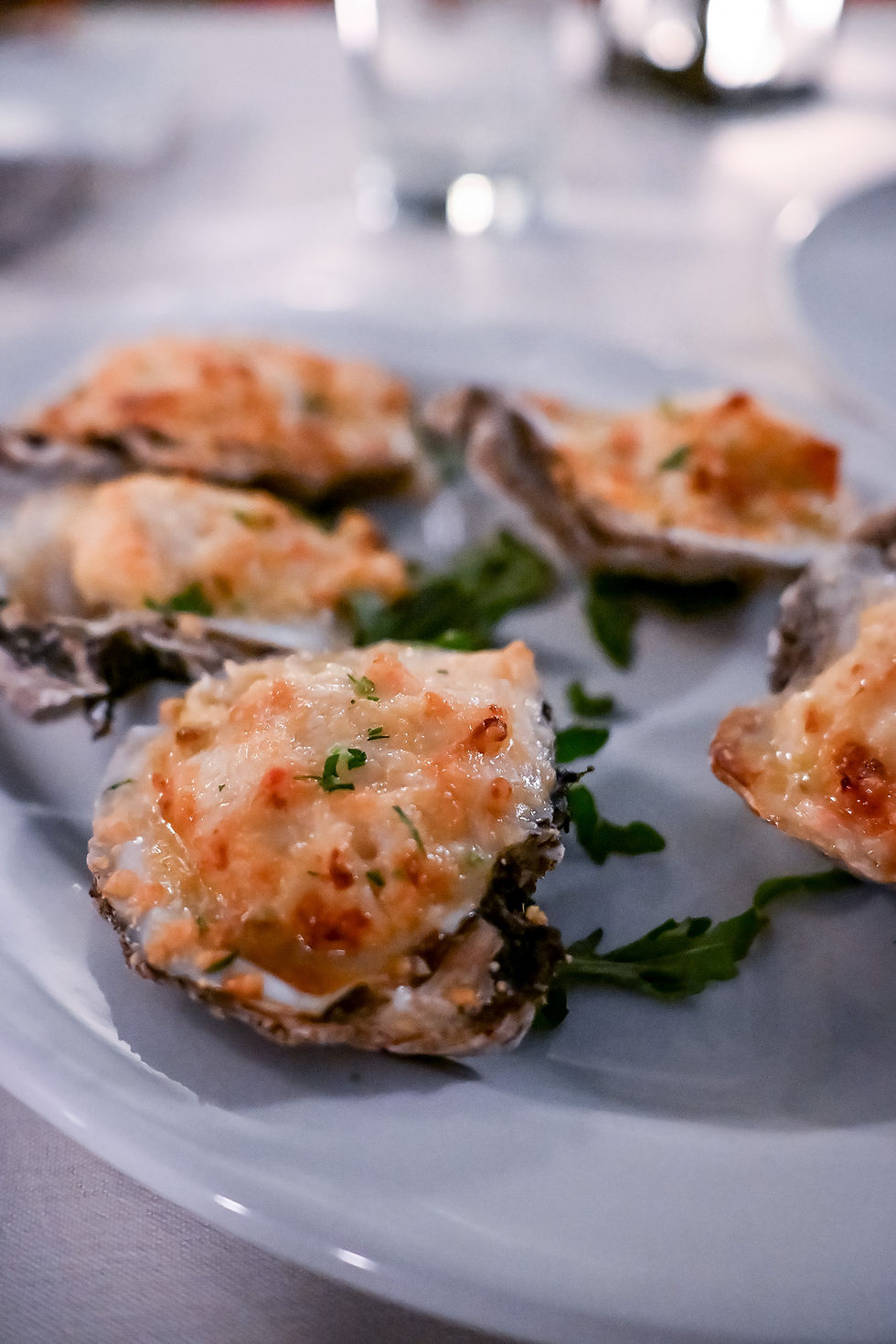 Cheese-topped oysters on a white plate garnished with greens, set on a white tablecloth in a dimly lit restaurant setting.