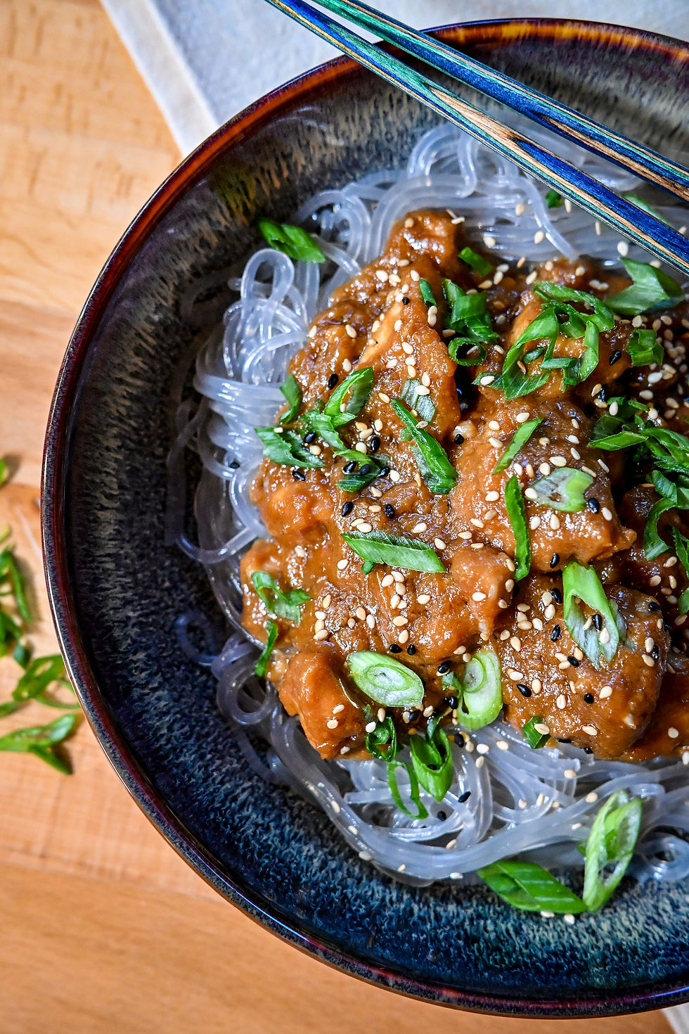 Close-up of a dark bowl of glass noodles topped with glazed meat, scallions, and sesame seeds, with blue chopsticks resting above.