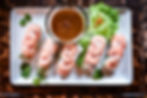 Shrimp spring rolls on a white plate with brown dipping sauce, lettuce, and cilantro on a dark wooden table.