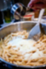 Hand sprinkles cheese from a metal cup onto cooked penne in a skillet, with a wooden spatula. Kitchen setting, warm mood.