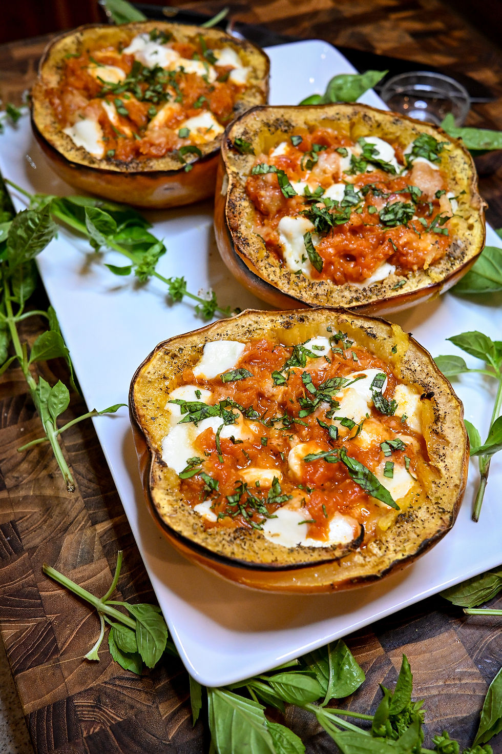 Three roasted stuffed squash halves topped with tomato sauce, melted cheese, and basil on a white plate with fresh herbs on wood.