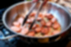 Sliced sausages are frying in a stainless steel pan on a stovetop. A hand with red nails uses tongs to turn them, with golden browning.