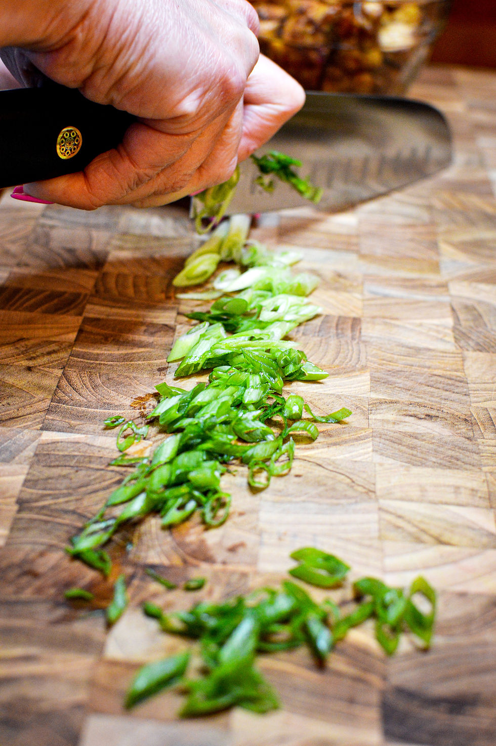 Hands chopping green onions on a wooden cutting board. The knife is mid-motion, scattering vibrant green slices across the surface.