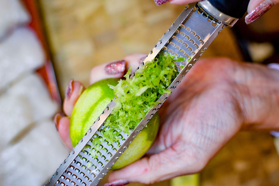 Hand with glittery nails zesting a green lime using a grater. Bright green zest collects on the metal surface. Warm background.