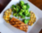 Grilled salmon with broccoli and pineapple slices on a white plate. The dish is set on a wooden table, creating a fresh, healthy vibe.
