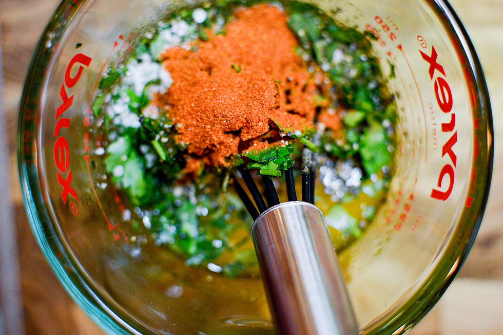 Mixing herbs, spices, and red powder in a glass measuring cup with the word "Pyrex" visible. A whisk is partially submerged.