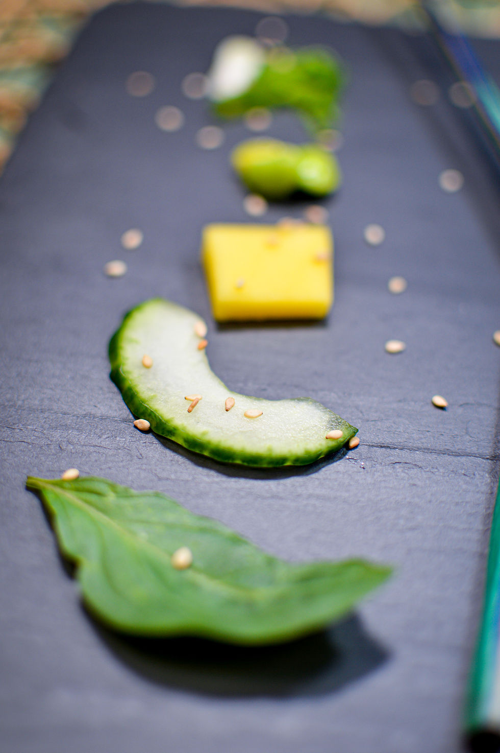Sliced cucumber, mango, and herbs with sesame seeds on a black slate. Bright colors create a fresh, vibrant mood. No text visible.