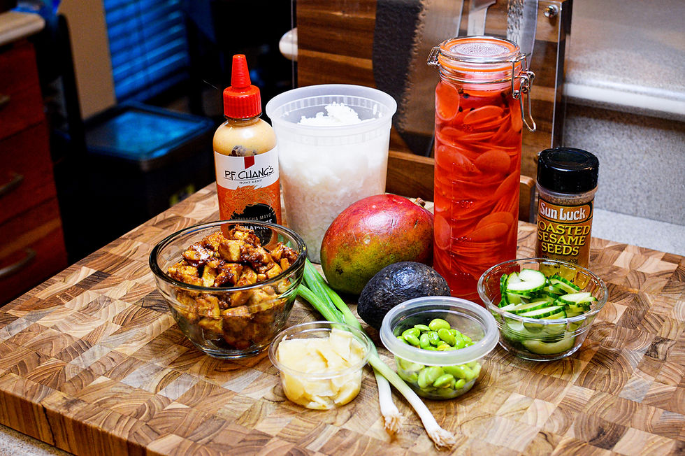 Ingredients on a wooden board: cubed chicken, mango, avocado, cucumbers, edamame, pickled carrots, ginger, scallions, sesame seeds, spicy mayo.