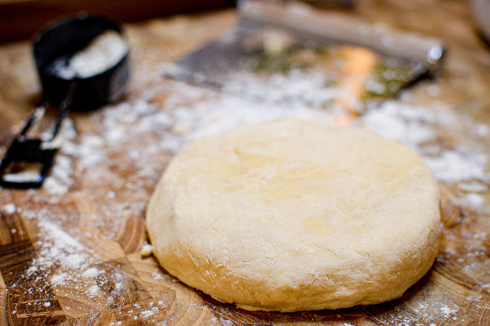Ball of dough on a floured wooden surface with a black measuring cup and a pastry cutter in the background. Rustic kitchen setting.