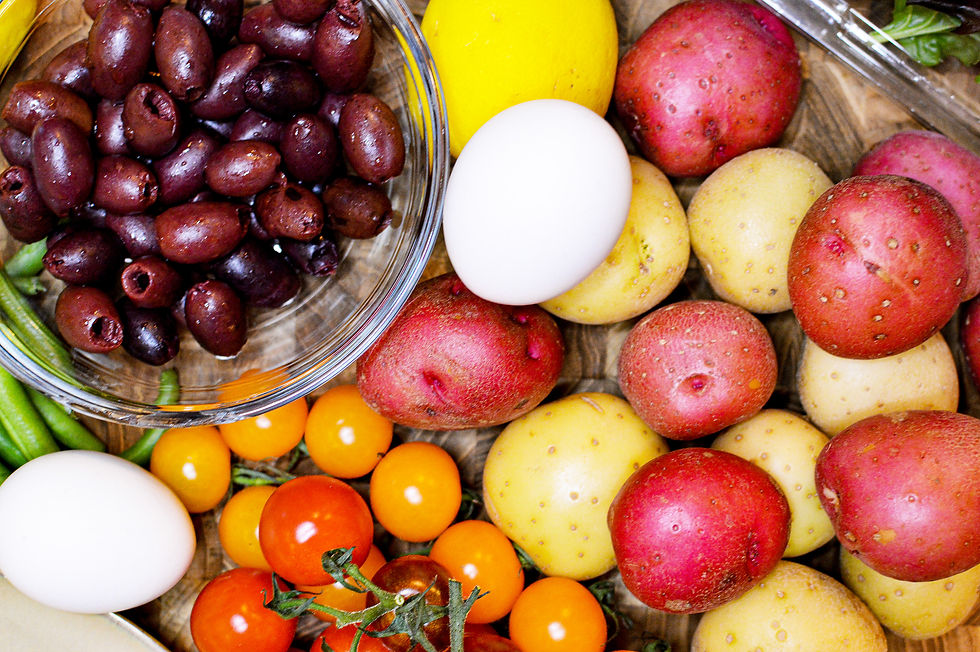 Bowl of dark olives, surrounded by colorful potatoes, cherry tomatoes, a lemon, and a white egg on a wooden surface. Bright and fresh.