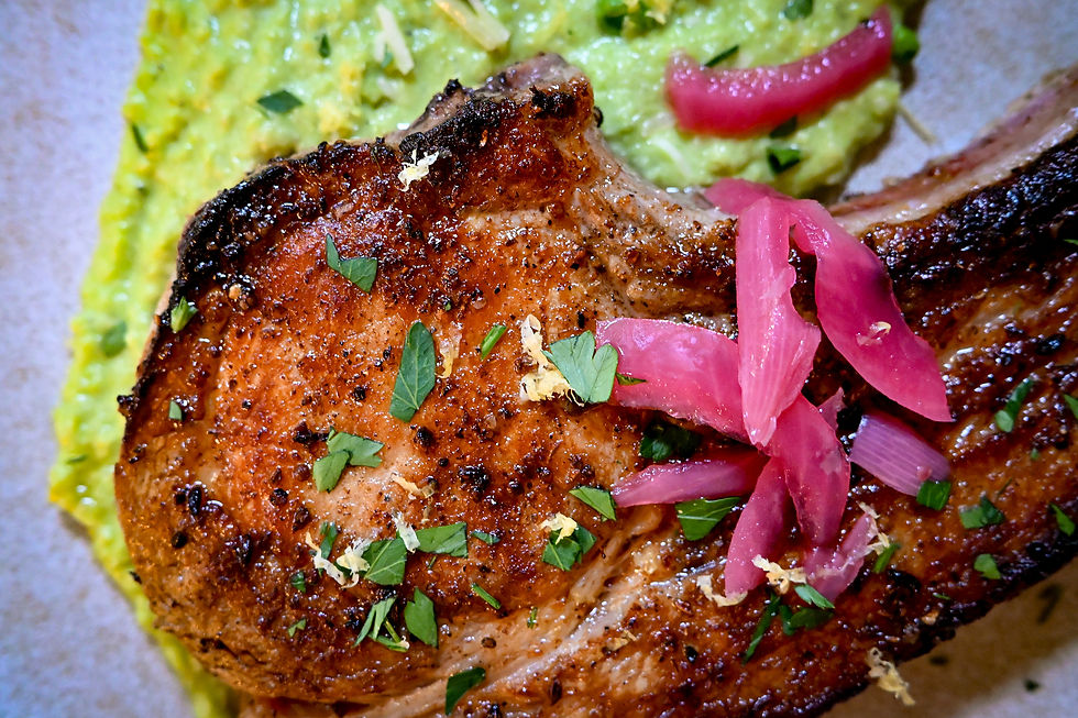 Close-up of a grilled pork chop topped with herbs and pickled onions, set against a backdrop of vibrant green mashed peas.