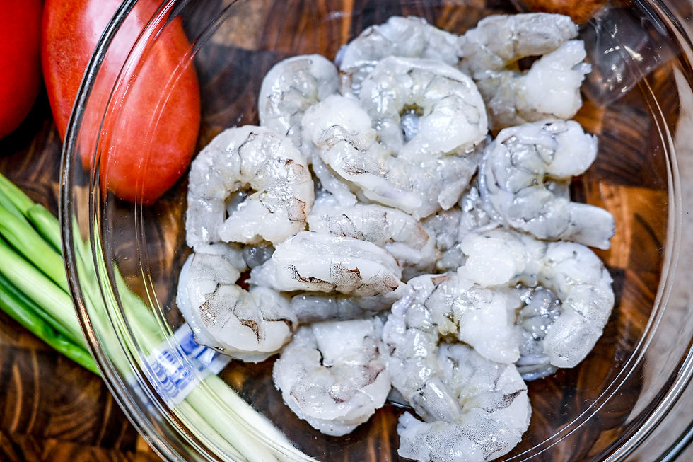 Raw shrimp in a clear bowl on a wooden surface, with whole tomatoes and green onions nearby. Fresh seafood preparation setting.