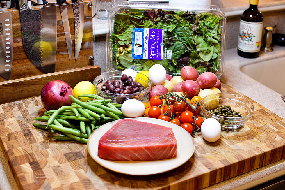 A wooden cutting board with fresh tuna, tomatoes, eggs, green beans, potatoes, and salad mix. Nearby, knives and a bottle. Vibrant colors.