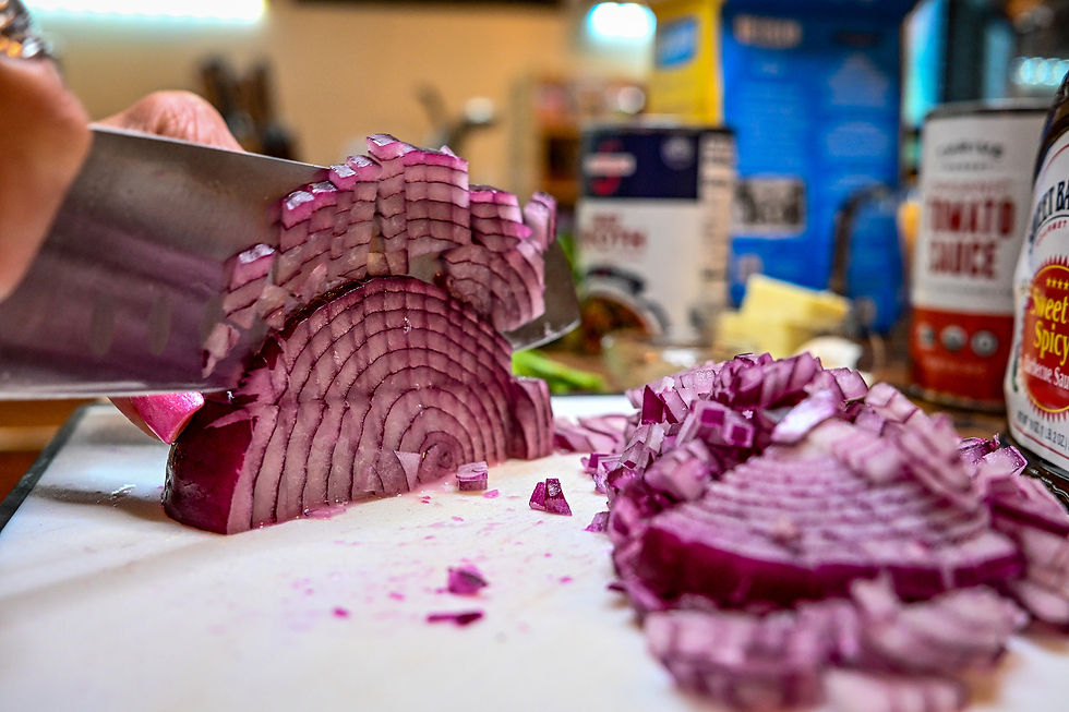 Close-up of a hand slicing a red onion on a cutting board, with chopped onion piles and sauce bottles in a kitchen.