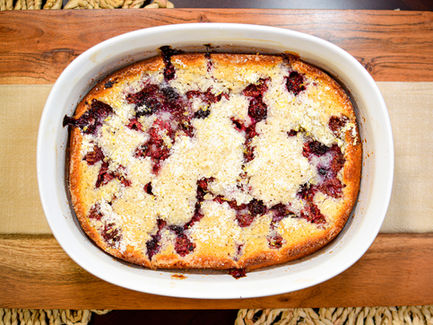 Golden-brown cobbler in a white dish, sprinkled with sugar, features baked red berries. Set on a wooden surface, evoking a homemade vibe.
