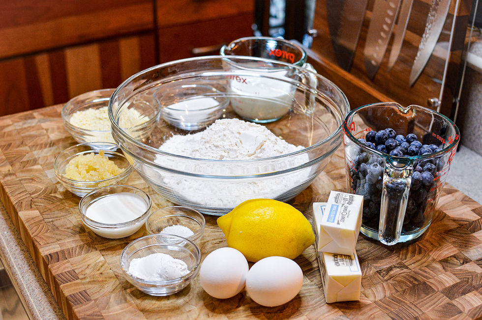 Baking ingredients on a wooden counter: flour, sugar, butter, blueberries, lemon, eggs, milk, in glass bowls and measuring cups.