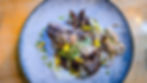 Grilled steak on a stone plate, topped with fried onions, green sauce, and yellow flowers, served on a wooden table.