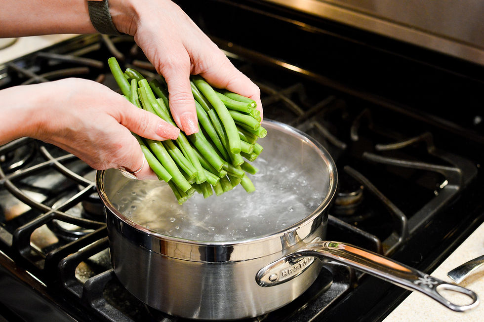 Hands holding fresh green beans over a pot of boiling water on a stove, preparing to cook. A stainless steel pot is on the black stove.