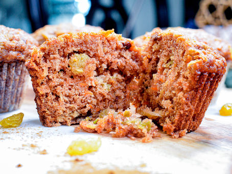 Close-up of a sliced carrot muffin with visible raisins on a wooden surface. Other whole muffins in the blurred background. Warm tones.