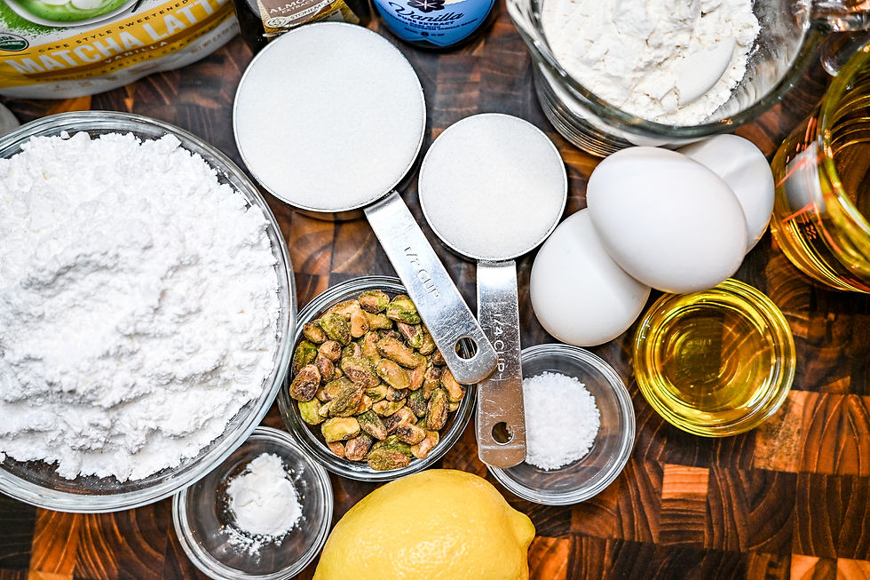 Ingredients on a wooden table: flour, sugar, eggs, pistachios, lemon, oil, baking soda, and a "Matcha Latte" label in the background.